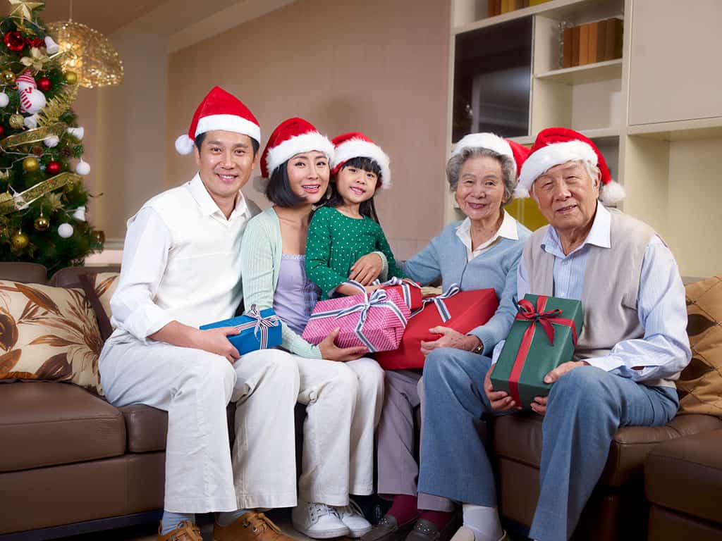 An Asian family sitting down on a couch with Christmas Santa hats on their heads while holding presents. This family consists of a young man and woman, one child, and an elderly woman and man. A Christmas tree can be seen in the background.