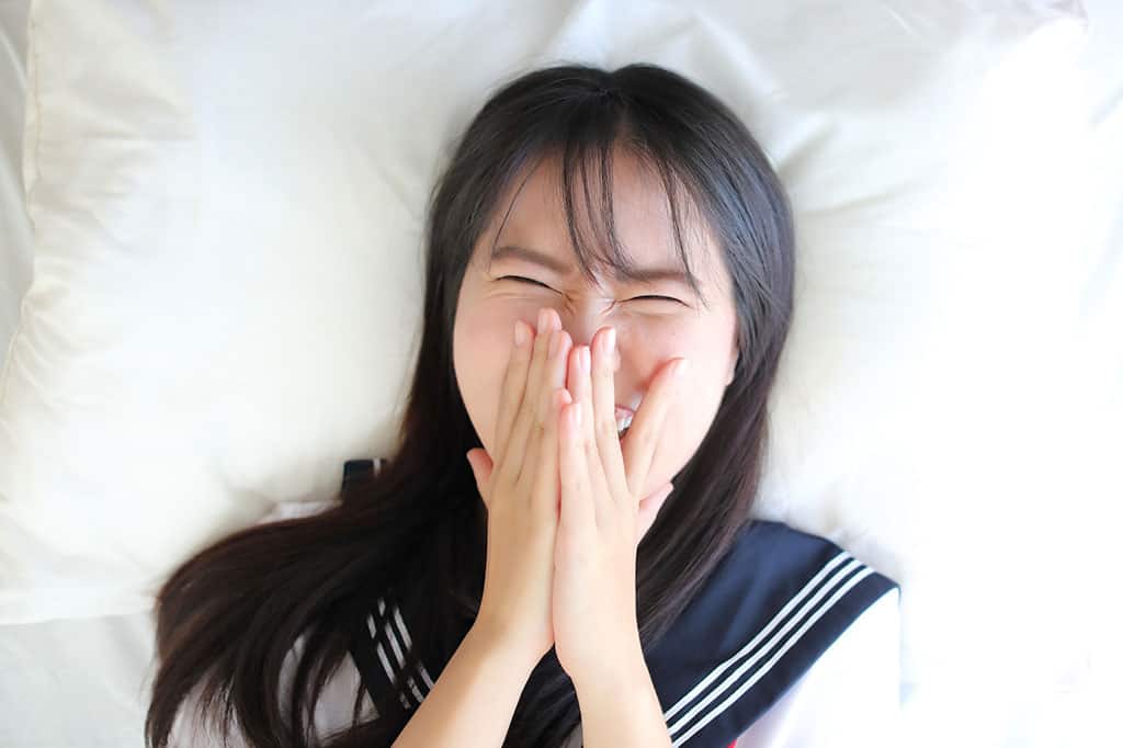 A young, Asian girl in a Japanese high school uniform lying down on a bed. Her hands are covering her mouth while she is laughing at something.