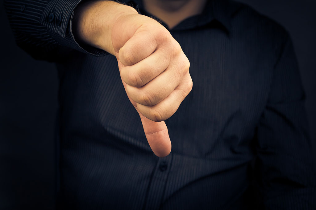 A close-up of a man’s hand giving a thumbs down. The man is wearing a button up black shirt with stripes which can be seen blurred in the background.