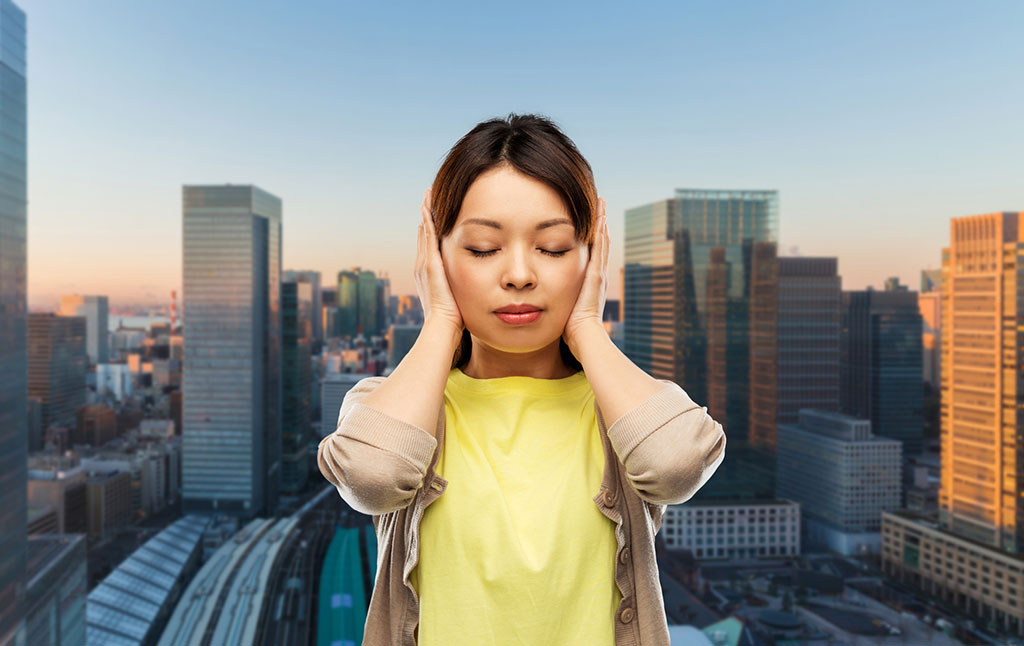 A young, Asian woman in the center of the image holding her hands up to her ears. In the background, the city (Tokyo) with many tall buildings can be seen.