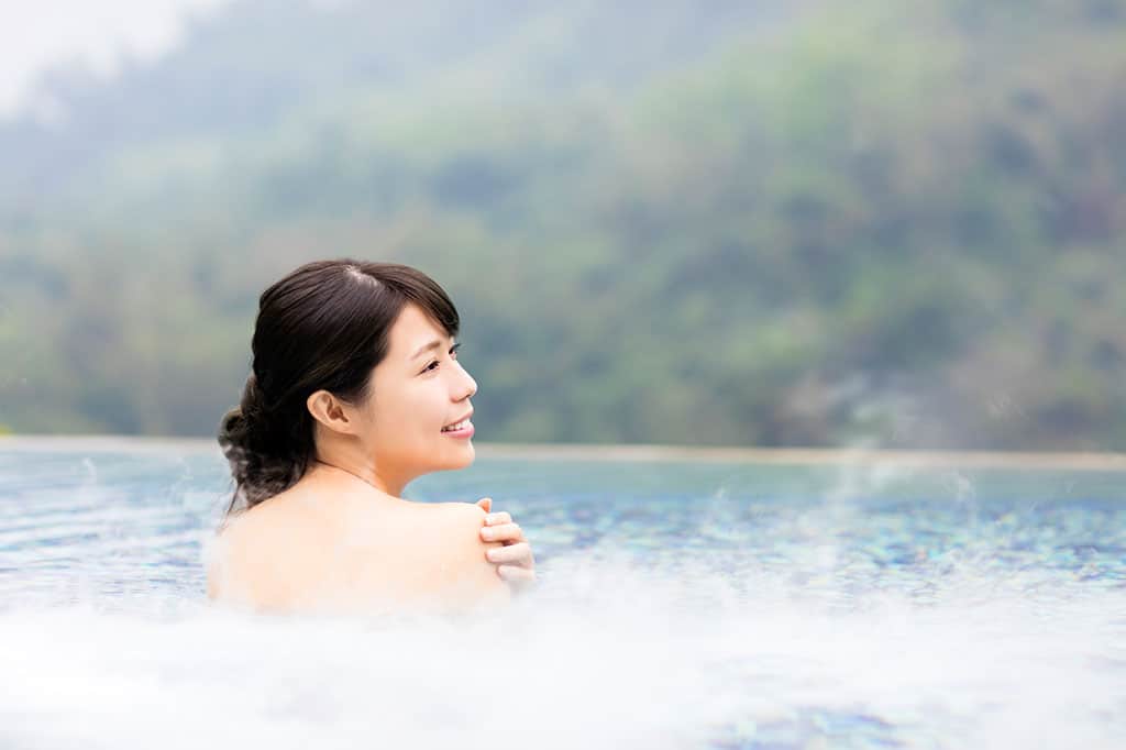 A young, Asian woman is relaxing in an outdoor hot spring, with trees and natured blurred in the background.