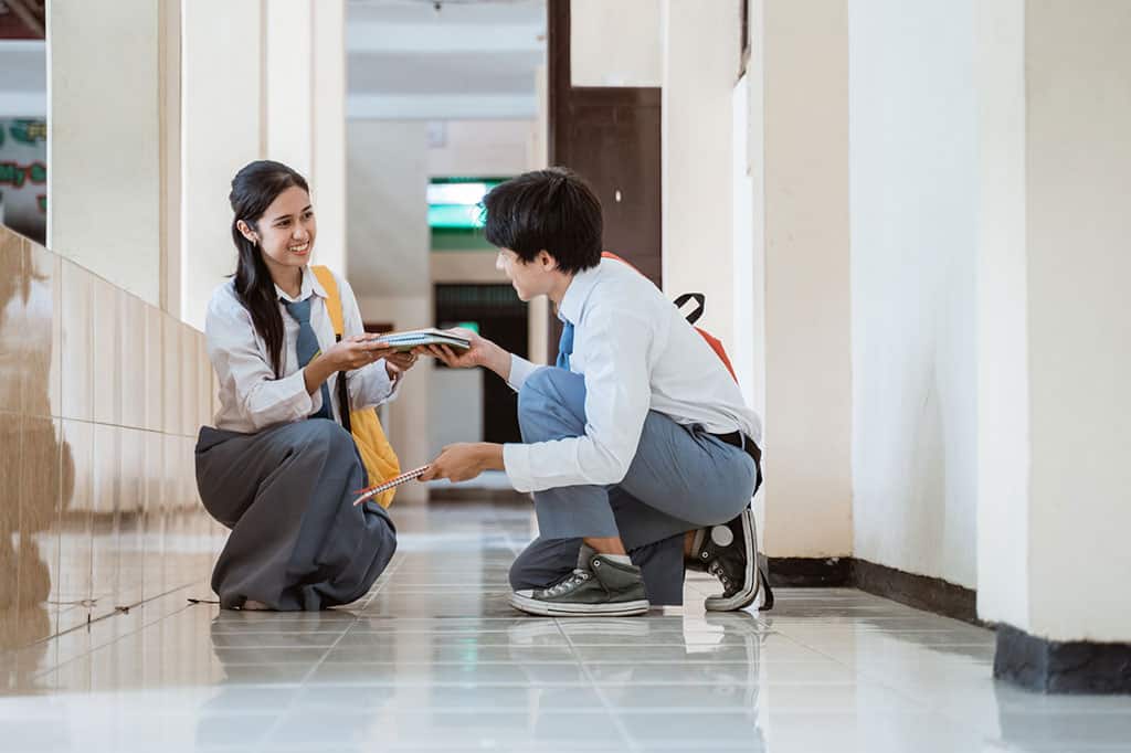 A young Asian boy crouching down and picking up books for a young girl, also crouching down and smiling at the boy. Both are wearing a school uniform (white dress shirt with a blue tie and grey skirt/pants).
