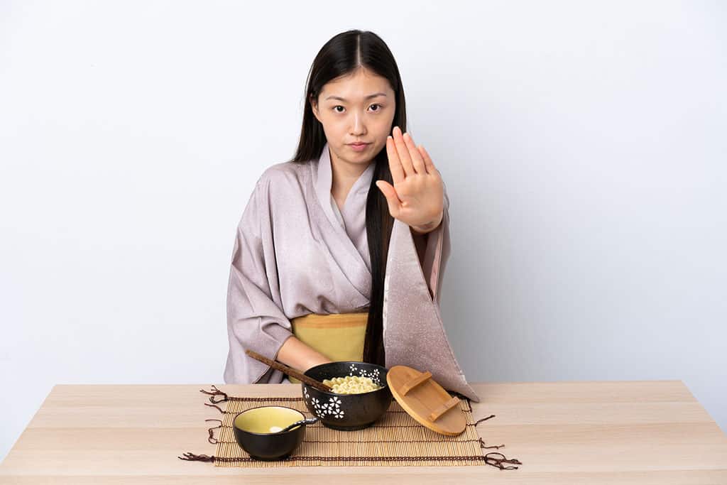A young Asian woman dressed in a Japanese style kimono holding up her left hand as if to say "stop." There are two black bowls on a table in front of her. One bowl is empty, and the other bigger bowl has ramen in it.