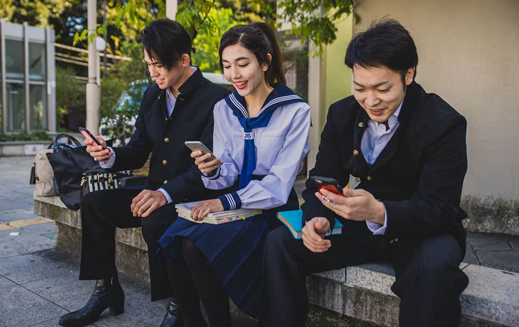 A young, Asian girl in a white, Japanese school uniform looking at her smartphone. She is sitting down in between two young Asian men, dressed in a Japanese school uniform. The men are also looking at their smartphones.