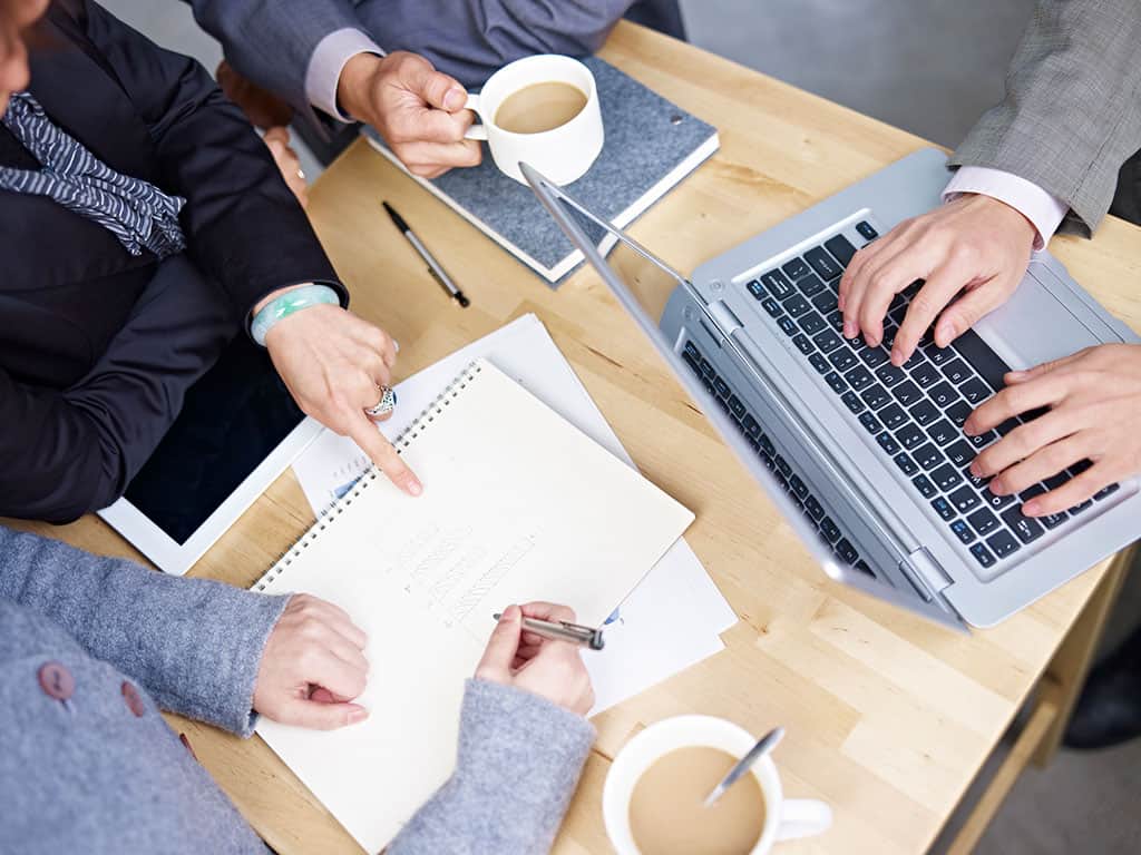 An overhead shot of 2 businessmen and 2 businesswomen sitting at a small wooden desk doing work. The two women are focusing on a notepad, while one man's right hand is holding a coffee, and the other man's hands are typing something on his silver laptop.