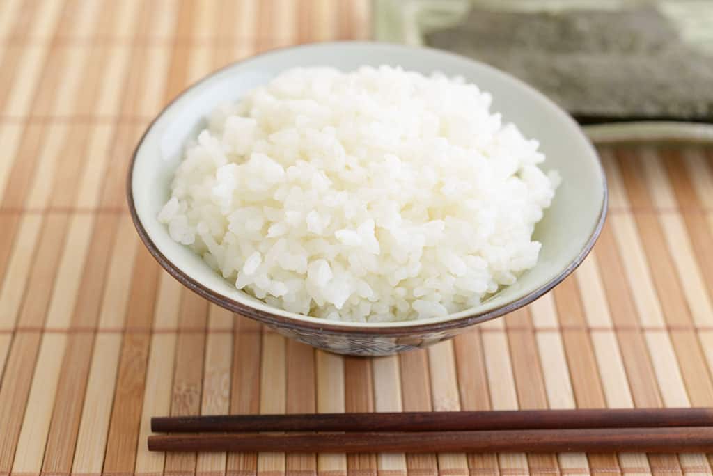 A bowl of cooked, white rice in a porcelain-like bowl on top of a wooden mat with a pair of brown chopsticks in front of it and nori blurred in the background.