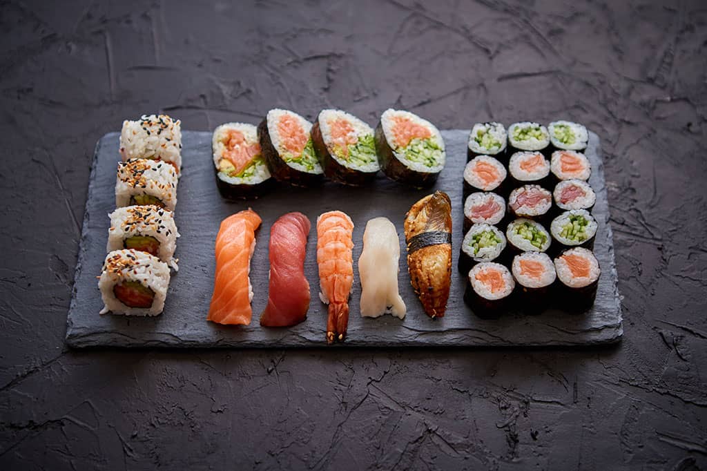 A flat, black plate on a black background. There are several types of nigiri and maki sushi on this plate (salmon, maguro, unagi, cucumber, shrimp, fish, etc.)