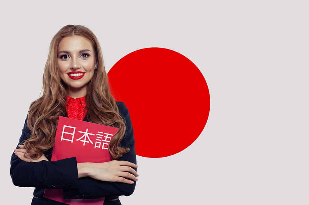 A young woman with brown hair is standing in front of the flag of Japan. She is smiling and holding a red book that has Japanese characters on it that translates to "Japanese."