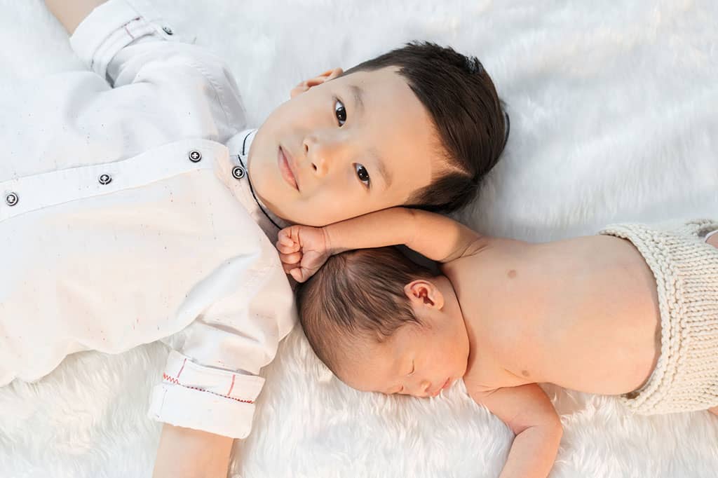 A photo of a young boy lying down on top of a white rug, with his feet extending past the left side of the picture. A baby boy is also lying on the rug, with his head next to the young boy’s, with his feet extending past the right side of the picture.