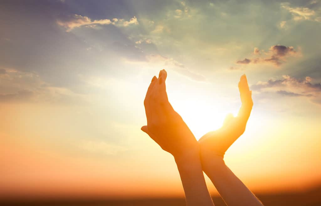 A woman’s hands are touching at the wrists to form a cup with her hands. She is holding her cupped hands up to the sky as if cupping the sun in her hands.