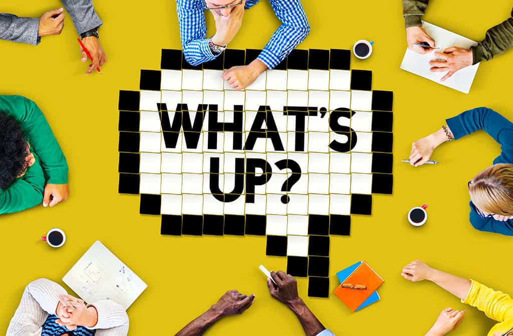 A birds-eye image of a group of 8 people sitting around a large, yellow, tabletop. Only the arms and hands of most people at the table can be seen. There are also cups of coffee, notebooks, and books on the table. In the center of the table is a white speech bubble with a black border, both of which are created by white or black colored squares. In this speech bubble, the words, “WHAT’S UP?” is displayed.