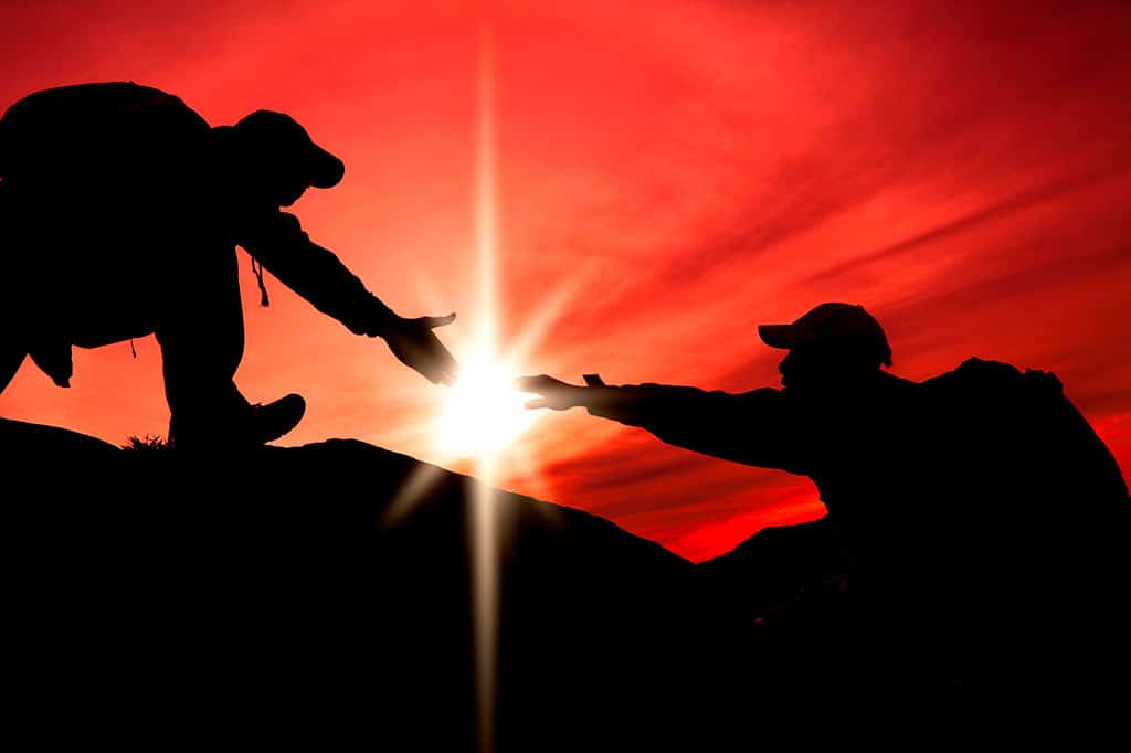 An image of two human silhouettes climbing up a hill. The person at the top is extending their hand to the outstretched hand of the person below. The shining sun is in the background, peering through space between the two people’s outstretched hands.