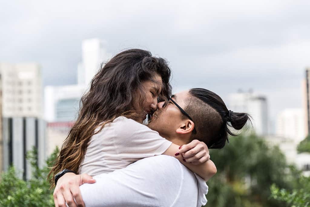 A young couple embracing one another. The woman on the left has her arms wrapped around the man’s neck, while the man to the right has his arms wrapped around her back. Their faces are close to each other as if about to kiss. Blurry buildings can be seen in the background.