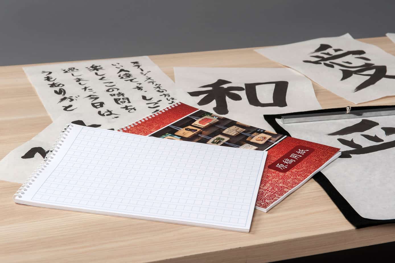 Pieces of paper on a desk that have Japanese kanji characters written on them with a brush and black ink. On top of these papers is a red notebook in the center on the image and a Japanese notebook for practicing writing characters is on top of it.
