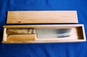 A nakiri vegetable knife with a walnut colored handle inside of a custom designed, tan box against a blue background.