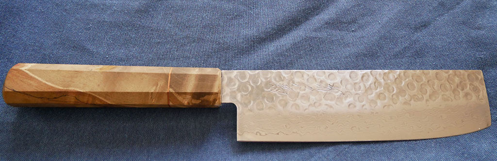 The nakiri Japanese vegetable knife laying on a blue cloth. This knife is from Oishya, and features a natural brown color wooden handle and silver blade with patterns on it.