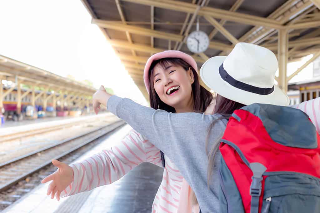 Two young women are standing on a platform at a train station. They are facing each other (with one woman’s back facing toward the camera), and have their arms open ready to embrace each other. The woman with her face towards the camera has a big smile, and looks extremely happy.