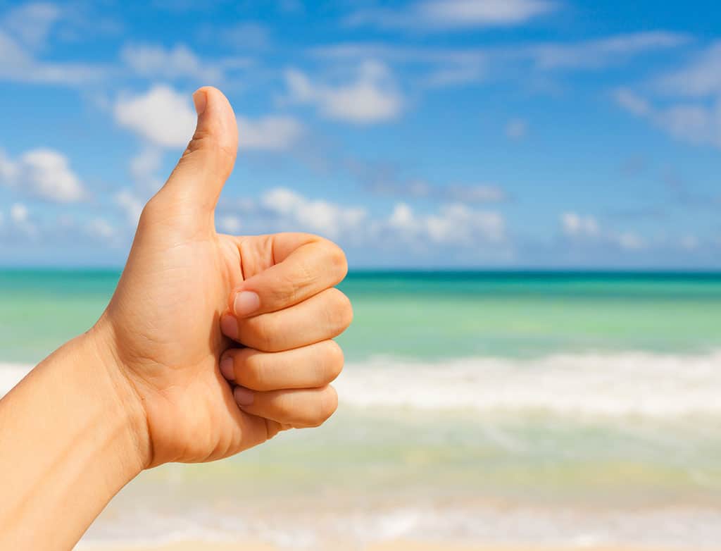 A close-up of someone’s left hand giving a “thumbs up” gesture. In the background, a beach with the blue sky and blue ocean can be seen.