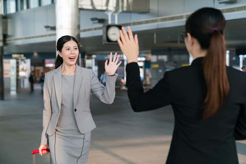 A woman wearing a gray business dress is walking while pulling a piece of luggage and is waving to another woman in the distance. The woman she is waving to is wearing a black business suit and has her back to the camera. This woman is waving back to the woman in gray.