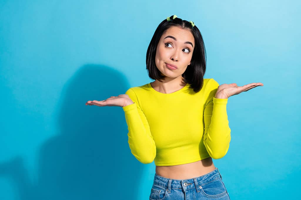 A young woman wearing a yellow-greeish top and blue jeans standing in front of a light blue background. She has both of her hands up (palms up) to her shoulder to gesture “I don’t know.”