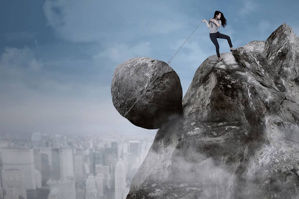 A woman is climbing up a mountain/rock while wearing blue jeans and a gray shirt. She is dragging a huge boulder up the mountain with a chain. The blue sky and a city with many tall buildings can be seen in the background.