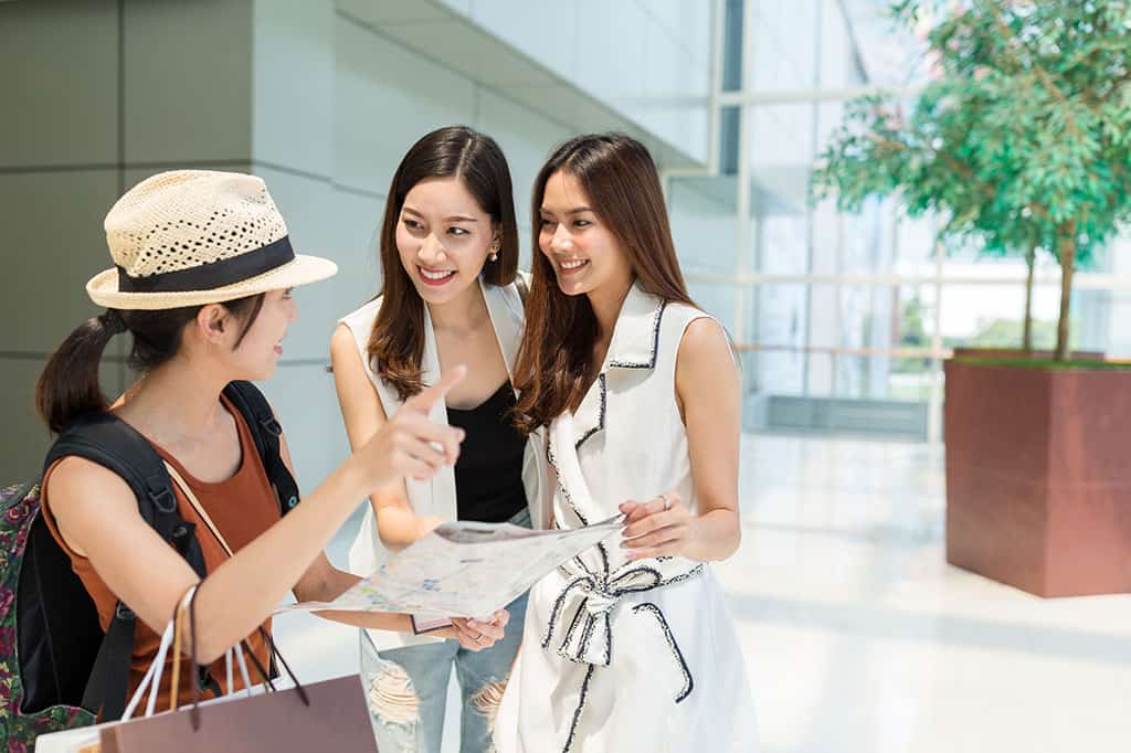 Three young women are studying next to each other. The woman on the left is holding a map and pointing to somewhere off in the distance. She appears to be asking for directions from the other two women. The two women on the right side of the photo are looking at the woman pointing and smiling at her.