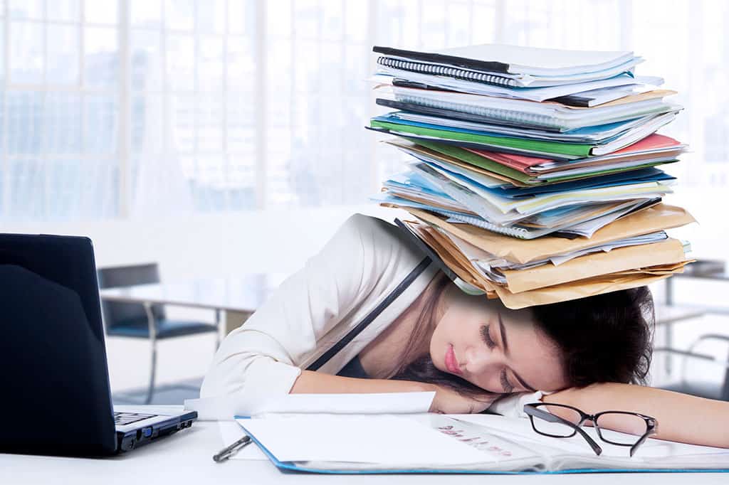 A young woman resting her head on her arm on top of a desk. On this desk, there is a black laptop next to her and an open binder in front of her. On the binder is a black pair of glasses and a black pen next to the binder. There is a lot of envelopes, folders, and notebooks that are all stacked up into one pile which is balanced on top of the woman’s face.