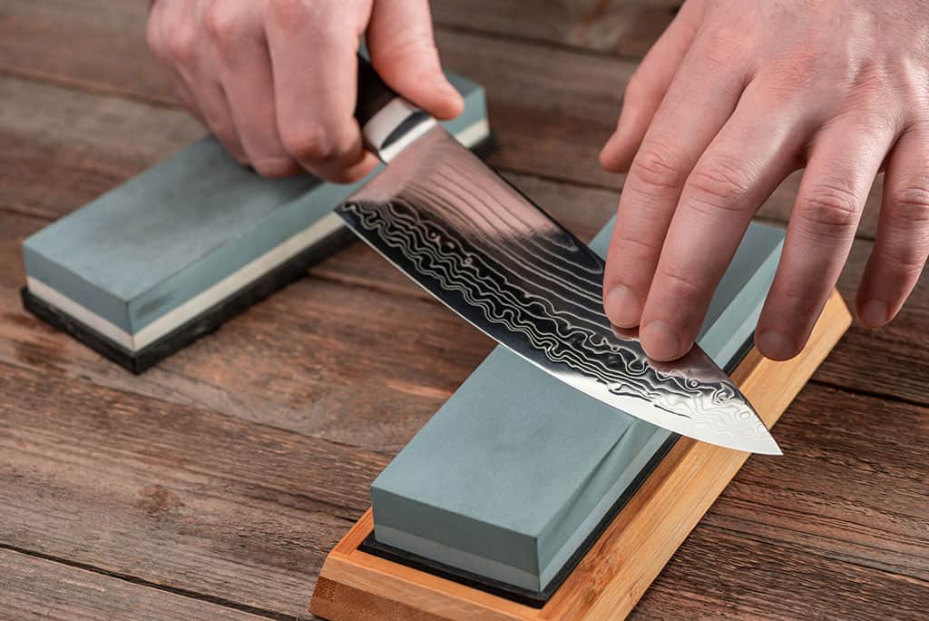 A close-up of a man’s hands holding a sharp gyuto style knife, sharpening it on a whetstone. There is another whetstone displayed in the background.