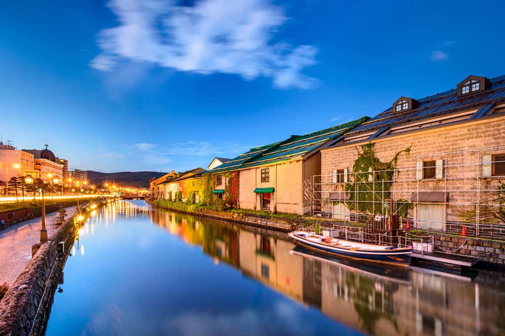 The Otaru Canal in Hokkaido.  On the left side of the canal/image there is a sidewalk running along the water, with buildings in the background.  On the right side, there are buildings running alongside the water.  An empty boat can be seen in the water. 