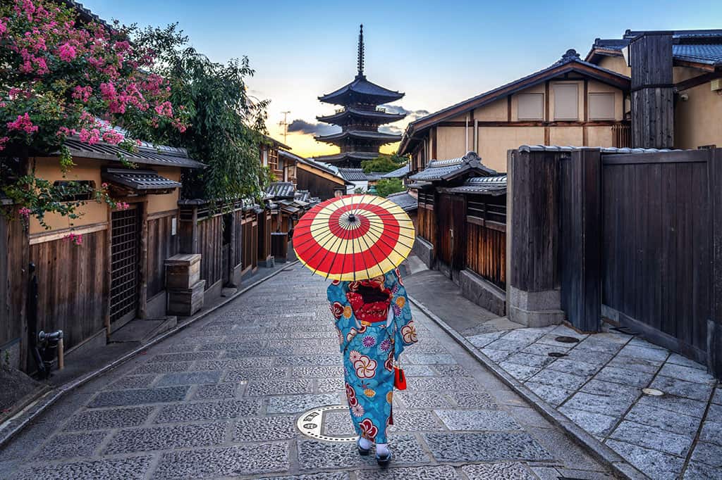 A woman dressed in a blue kimono/yukata holding an umbrella with a red, spiral design on it. She is walking through some buildings with a Japanese pagoda in front of her.