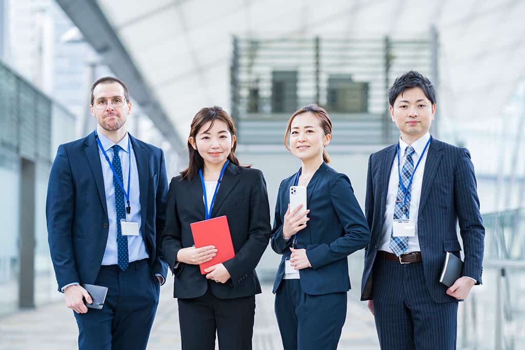 Four people (2 men on the left and right side and two women in the middle of them) are wearing business suits and posing next to each other.  2 of the men and 1 woman are holding small books, like a planner, and 1 woman is holding up her smartphone.  